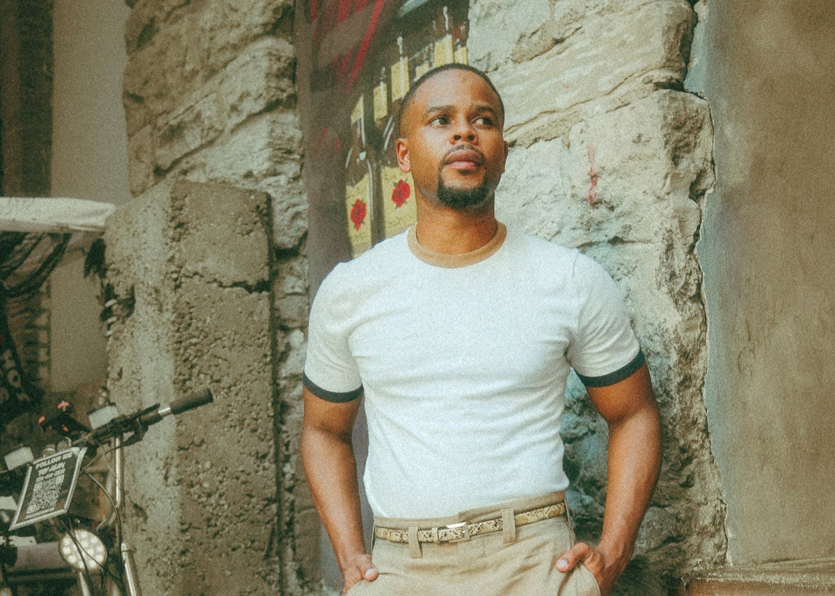 Man in a white t-shirt and beige pants standing against a stone wall with a bicycle in the background.